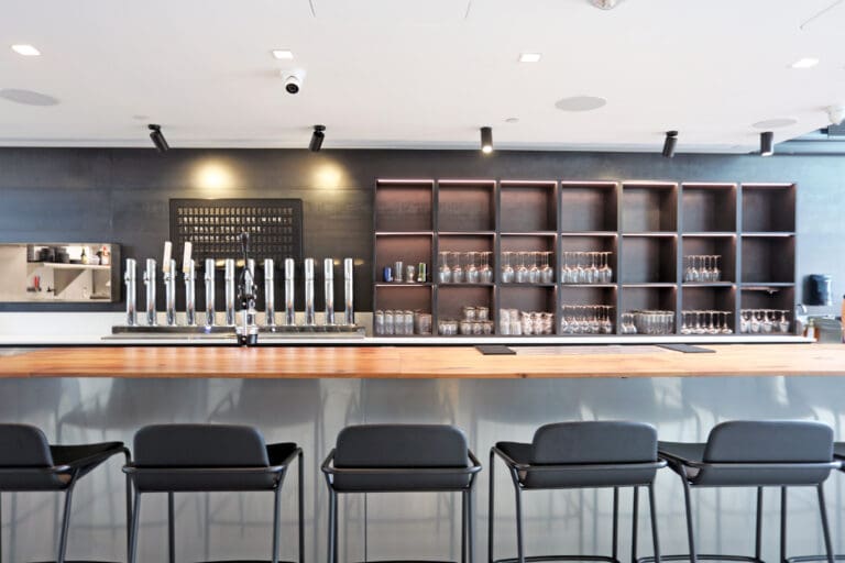 A modern bar with a long wooden counter, several black chairs, numerous taps for dispensing drinks, and shelves filled with various glassware creates the perfect ambiance for enjoying a K2 Electric cocktail.
