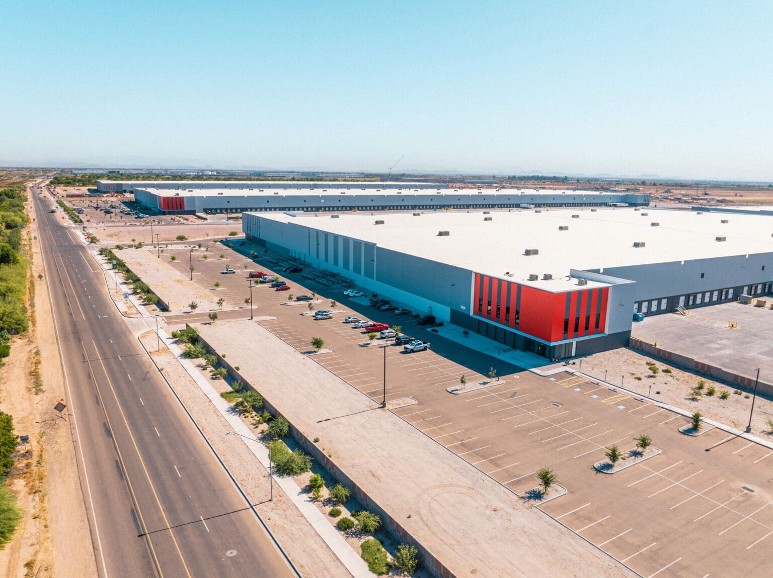 Aerial view of large industrial warehouses with orange accents, adjacent parking lots, and a K2 Electric service road beside them under a clear blue sky.