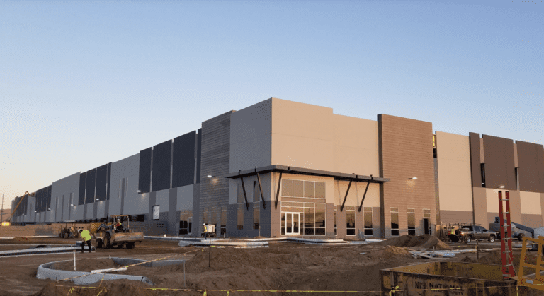 A large industrial warehouse under construction, with workers and machinery from K2 Electric on site, partially completed exterior walls, and a clear sky in the background.
