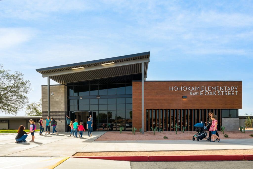 People are gathered in front of Hohokam Elementary at 8451 E Oak Street. The modern elementary school features a sleek design with large windows and wooden accents.