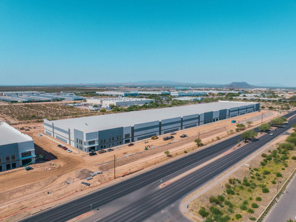 Aerial view of a large industrial warehouse under construction in Tucson, with an adjacent road and parked vehicles on a clear day. This future eCommerce center promises to transform the local landscape.