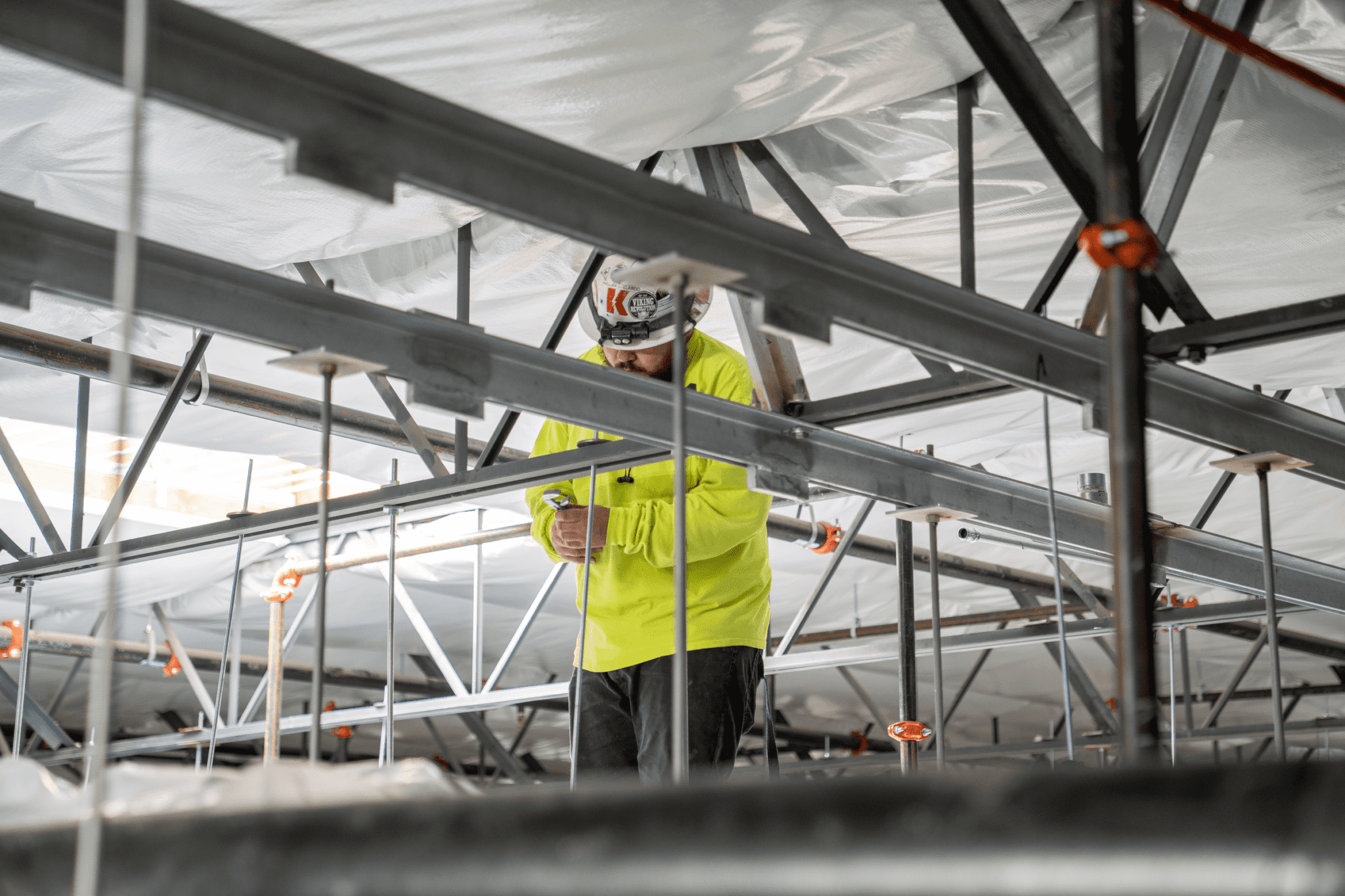 A construction worker wearing a high-visibility jacket and a helmet stands on scaffolding, utilizing a K2 Electric tool while surrounded by a framework of beams and supports.