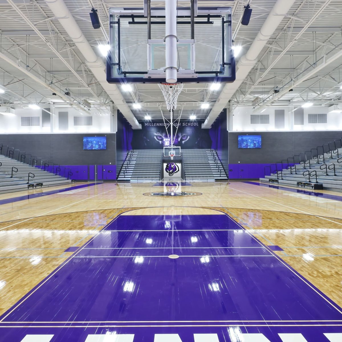 Empty indoor basketball court with purple markings, bleachers on both sides, and bright overhead lighting at Millennium High School.