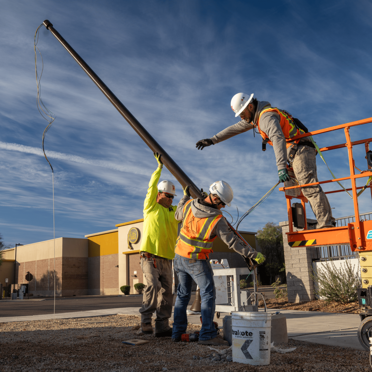 Three construction workers in safety gear install a utility pole using a hydraulic lift outdoors on a clear day near Bullard Wash.