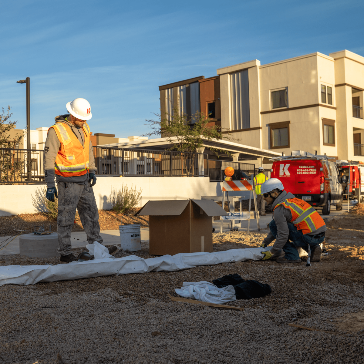 Two construction workers, one standing and one kneeling, work with materials on a gravelly site near modern buildings along Bullard Wash. Both wear safety vests and hard hats. A red construction vehicle is visible in the background.