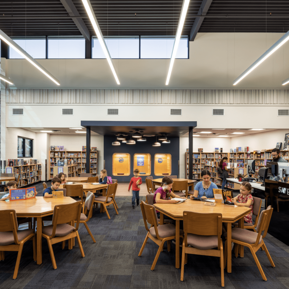 A group of people, including children and adults, are reading and working at tables in a modern, well-lit library with large windows and bookshelves at the Hohokam Elementary School.
