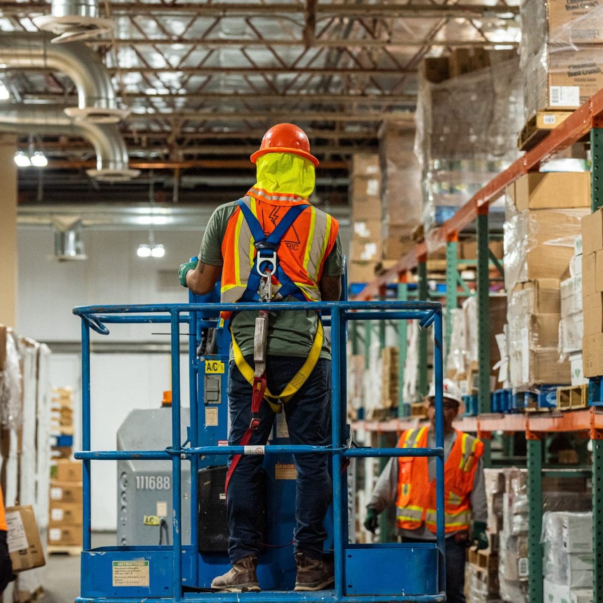 Workers in safety gear operate a lift in a warehouse aisle filled with stacked boxes and shelves, reminiscent of the organized systems that keep the Phoenix Unified School District running efficiently.