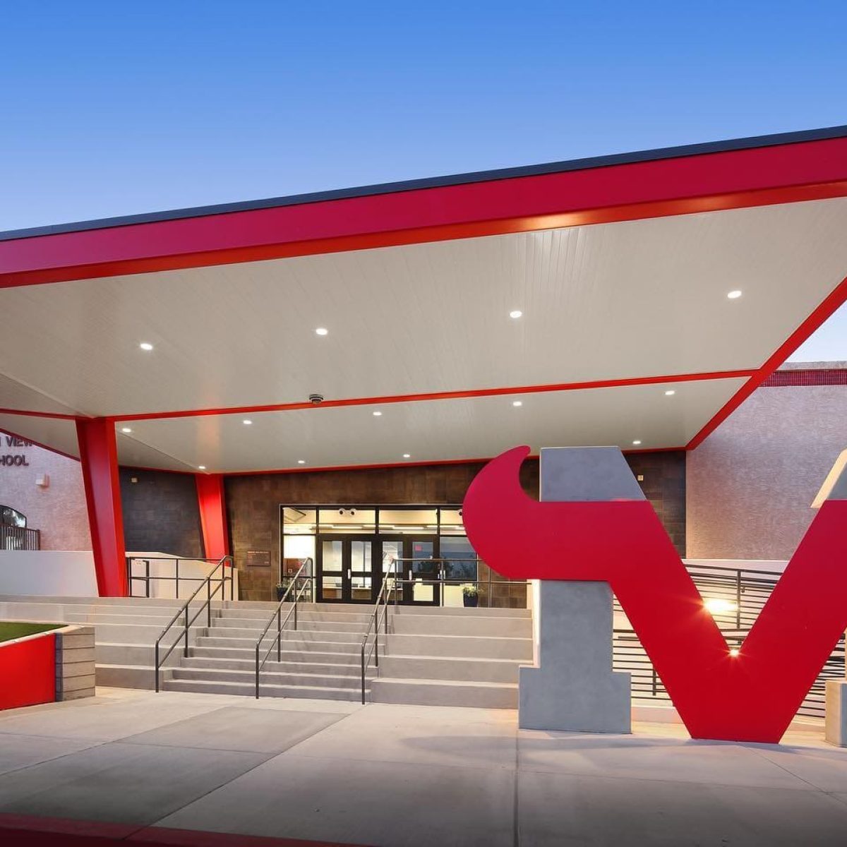 The entrance of Mountain View High School features modern architecture with red accents, a large M-shaped bullhorn sculpture, and a well-lit foyer, making it an impressive sight.