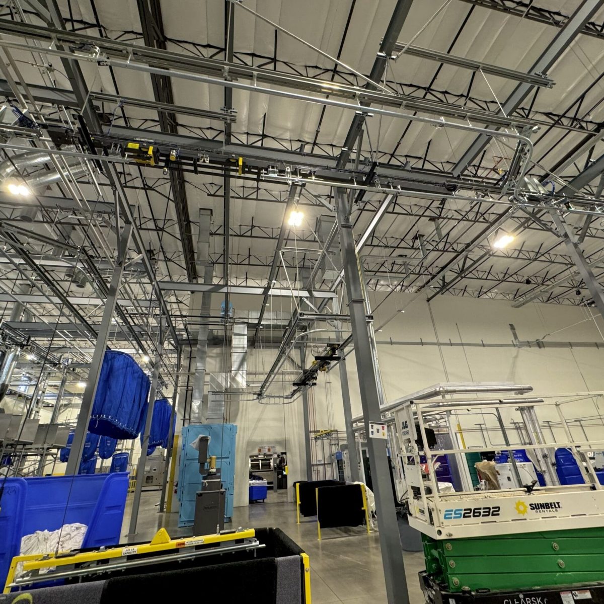 Interior image of a large warehouse with industrial machinery, overhead conveyor belts, and storage containers. Ceiling lights illuminate the area.
